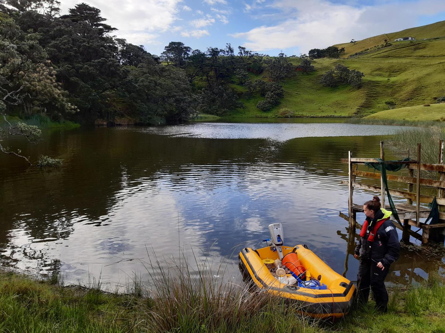 A rural setting and a large pond. On the pond is an inflatable boat that is about to be boarded by a person wearing a life jacket. The boat is filled with scientific equipment.